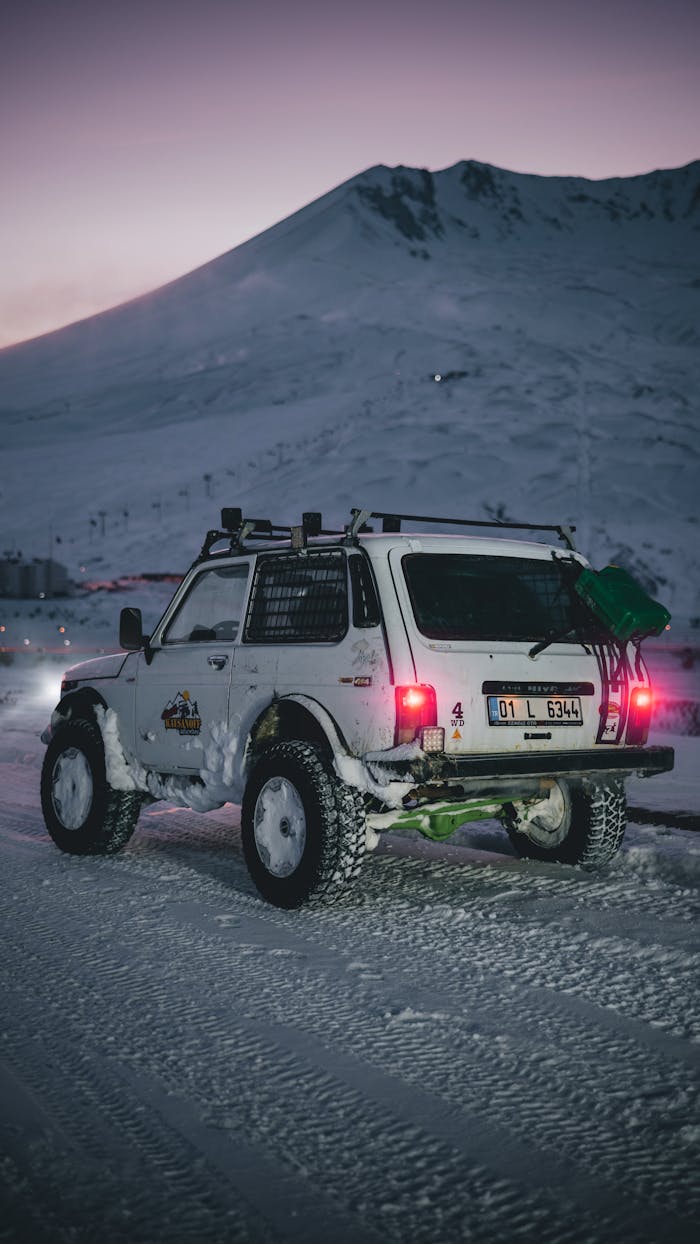 A rugged white 4x4 vehicle traverses a snowy mountain road at twilight.