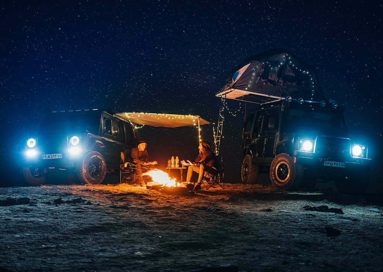 Two campers enjoying a campfire between jeeps with a starry sky backdrop in Razavi Khorasan, Iran.
